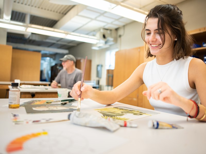 A woman sitting at a desk with different paints and pencils, working on a unseen drawing