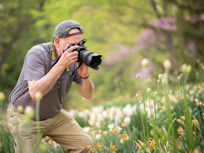 A photographer crouching down in a vibrant garden and taking a closeup photo of some flowers